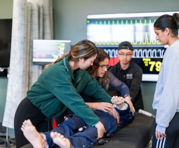 Three students are shown learning a bed-side procedure from a faculty member.
