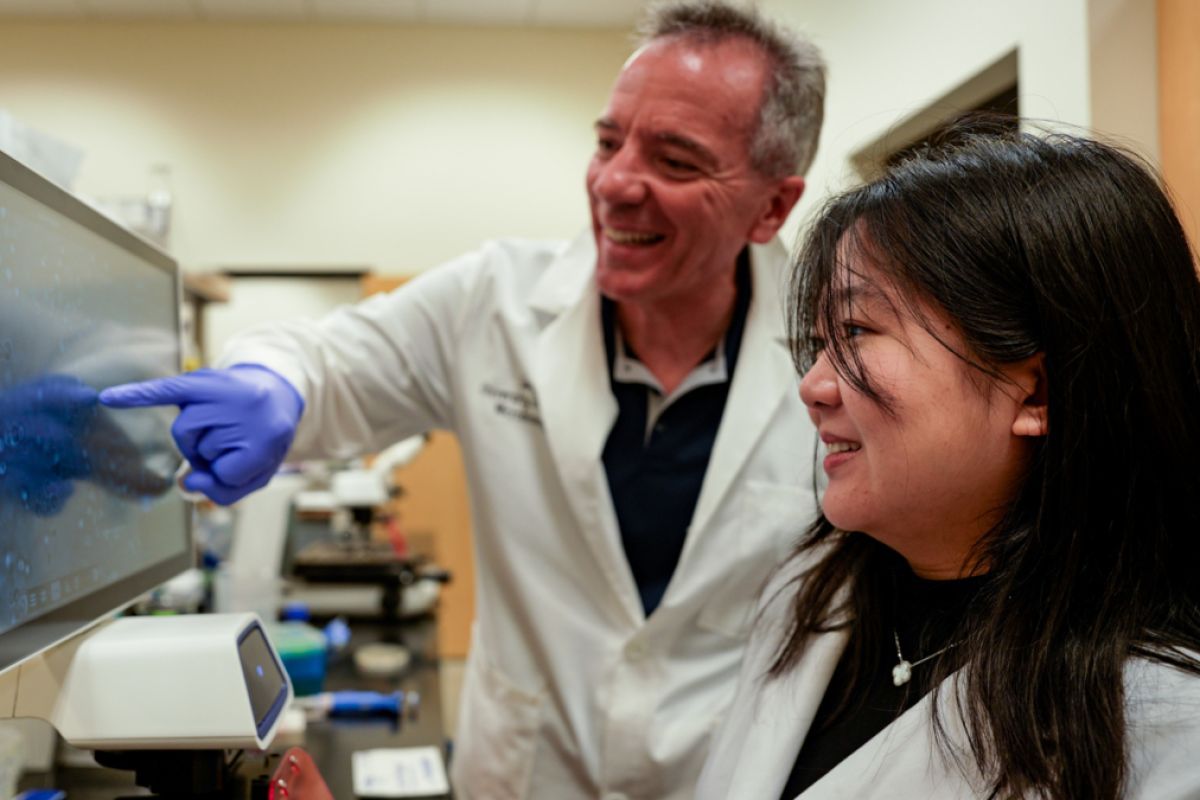a student wearing a lab coat looks at a computer with a professor
