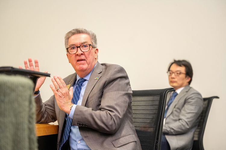 A man in a grey suit jacket and blue button shirt and tie sits and talks with his hands while another person sits behind him listening. 