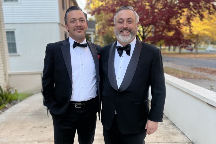 Two brothers stand in an outdoor setting wearing nice suits smiling together. 