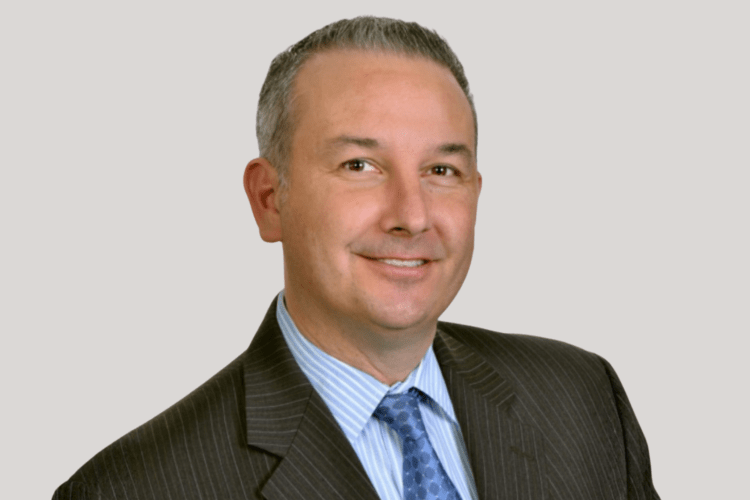 A headshot portrait of a man wearing a grey suit jacket a blue tie and a light blue under shirt. He has short gray hair and brown eyes.  
