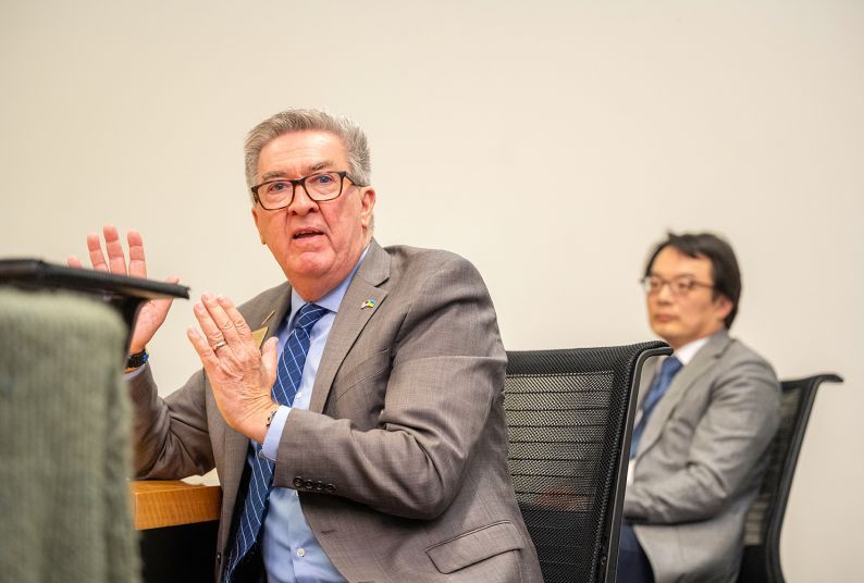 A man in a grey suit jacket and blue button shirt and tie sits and talks with his hands while another person sits behind him listening. 