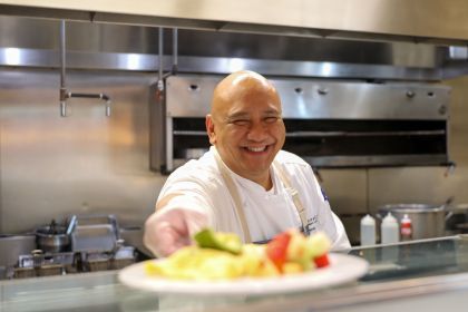 A man in a white chef coat smiles while handing a plate of food across the counter.